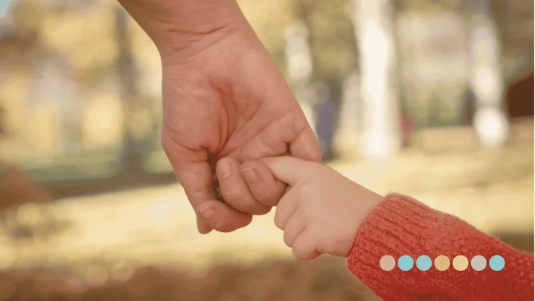 Krupni plan odrasle ruke koja nežno drži malu dečiju ruku u narandžastom rukavu, simbolizujući podršku porodici i detetu sa autizmom.